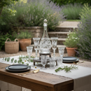 Dining table set with glasses, a decanter, and plates in a garden setting.