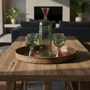 Decorative setup on a wooden table with a bottle, glasses, and a tray in a room with plants and furniture.