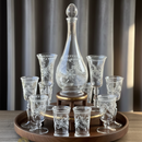 Set of etched glass decanter and glasses on a wooden tray with a neutral background.