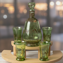 Green glass decanter set with four glasses on a wooden stand against a blurred background.