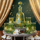 Green glass decanter and glasses with floral designs on a wooden stand, surrounded by white flowers.