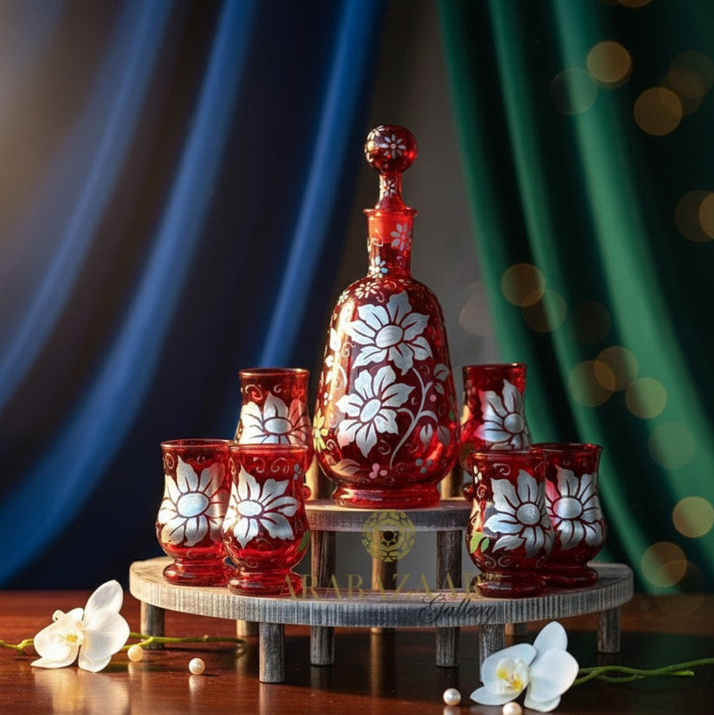 Set of red glass decanter and tumblers with floral designs on a wooden stand, against a blurred curtain background.