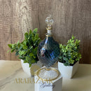 Decorative glass bottle with gold accents on a marble surface with plants and 'Arabazza Gallery' branding.