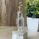 Decorative glass bottle with floral etching on a white pedestal, with a plant in the background.