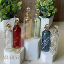 A collection of handblown glass perfume bottles with 14K gold trim, displayed on white pedestals. The bottles are clear and colored, featuring intricate etched designs.
