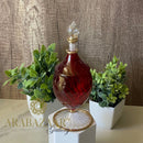 Decorative bottle with red liquid on a white stand, surrounded by plants against a wooden background.