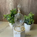 Decorative glass bottle with gold accents on a white pedestal, surrounded by green plants against a wooden background.