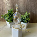 Decorative glass bottle with gold accents on a white pedestal, flanked by potted plants against a wooden background.